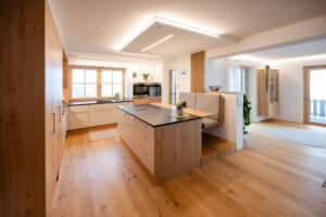 Eine offene Küche mit Plattform‑Frühstücksbar bzw. Essnische mit schwebender gepolsterter Bank, und gesenkter Decke mit integriertem Lichtelement – elegant durchdacht von Tischlerei Scharnreitner.