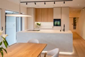 Eine moderne, offene Wohnküche mit beigen grifflosen Fronten, Holzakzenten in Form eines Oberschranks, Essbereich und einer Kochinsel in passender Beton bzw. Steinoptik in minimalistischem Design von Tischlerei Scharnreitner.