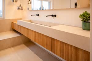 Ein modernes Badezimmer mit großformatigen Bodenfliesen, Sitz- und Ablagefläche und einem schwebendem Doppelwaschtisch in zusammenpassender beiger Steinoptik, schwarzen wandmontierten Armaturen, Pendelleuchte, Waschtischunterbau aus Holz mit moderner geriffelter Oberfläche und einem Spiegelschrank von Tischlerei Scharnreitner.