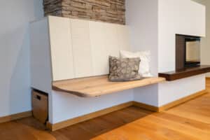 Eine gemütliche Stube mit Kamin, Holz‑Sitzfläche mit Kermaik-Lehne, und einer versteckten Nische mit Holzkiste als raffinierte Aufbewahrungslösung von Tischlerei Scharnreitner.