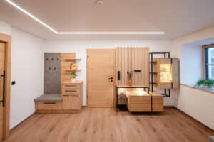 Eine moderne Wohnwand-Kombination mit Vitrine einem modernen Sprossenpaneel als Rückwand und mattschwarzem Eisenrahmen und Ablageflächen, gegenüber eine Garderobe mit grauer gepolsterter Sitzfläche von Tischlerei Scharnreitner.