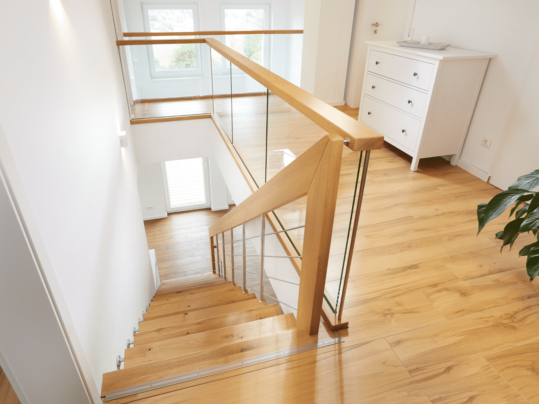 Treppenmeister zeigt ein lichtdurchflutetes Vorzimmer mit einer Galerie und einer weissen Kommode mit hellen Holztreppen als Zugang in das untere Stockwerk.