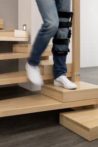 Person mit Beinverletzung benutzt eine Holztreppe mit alters- und beeinträchtigungsgerechter zusätzlicher Halbstufe für besseren Treppenauftritt.