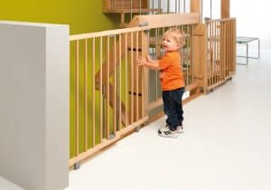 Lachendes Kleinkind steht hinter der Kindersicherung der Holztreppe im Obergeschoß eines Hauses.