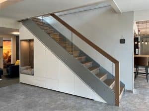 Eisentreppe im Industrialstil mit dunklem Holzhandlauf, Glasgeländer, Holzstufen und einem Treppenschrank mit grifflosen Fronten in Weiß, einer Garderobennische mit Sitzgelegenheit und jeder Menge Stauraum.