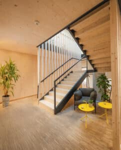 Ein großzügiger Holztreppen‑Aufgang mit Glasgeländer, Geländerahmen, Handlauf und Wangen aus mattschwarzem Stahl und einem umliegendem Wartebereich von Treppenmeister.