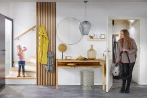 Ein Kleinkind steht am Treppenende und lacht seiner Mutter zu, die mit dem Staubsauger aus der Abstellkammer kommt, welche durch einen Wandverbau und durch die Nutzung des Untertreppenstauraums von Treppenmeister erschaffen wurde, die Länge des Wandverbaus bietet genügend Platz für einen Konsolentisch, einem großen Wandspiegel, einem schwebenden Wandregal und einem integrierten Holzsprossen-Wandpaneel als Garderobenaufhängung.