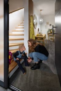 Mutter und Kind am Ende einer Treppe mit beleuchtetem Holzhandlauf, integriertem Holzsprossen-Wandelement welches als Garderobe dient und einem Wandverbau der als eine Extra-Abstellkammer funktioniert von Treppenmeister.