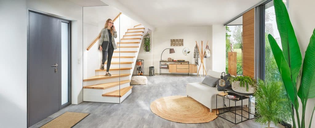 Eine Frau schreitet über ihre Treppe mit Holzhandlauf, Stufen mit Holzauftritt, freiem Untertreppenstauraum und einer deckenhohen Glaswand am Treppenende von Treppenmeister.