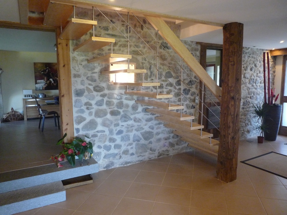 Eine Kragarmholztreppe mit vertikalen Eisenstäben als Geländer, hellem Holzhandlauf und passenden schwebenden Holztreppen befestigt an einer rustikalen Steinwand von Treppenmeister.
