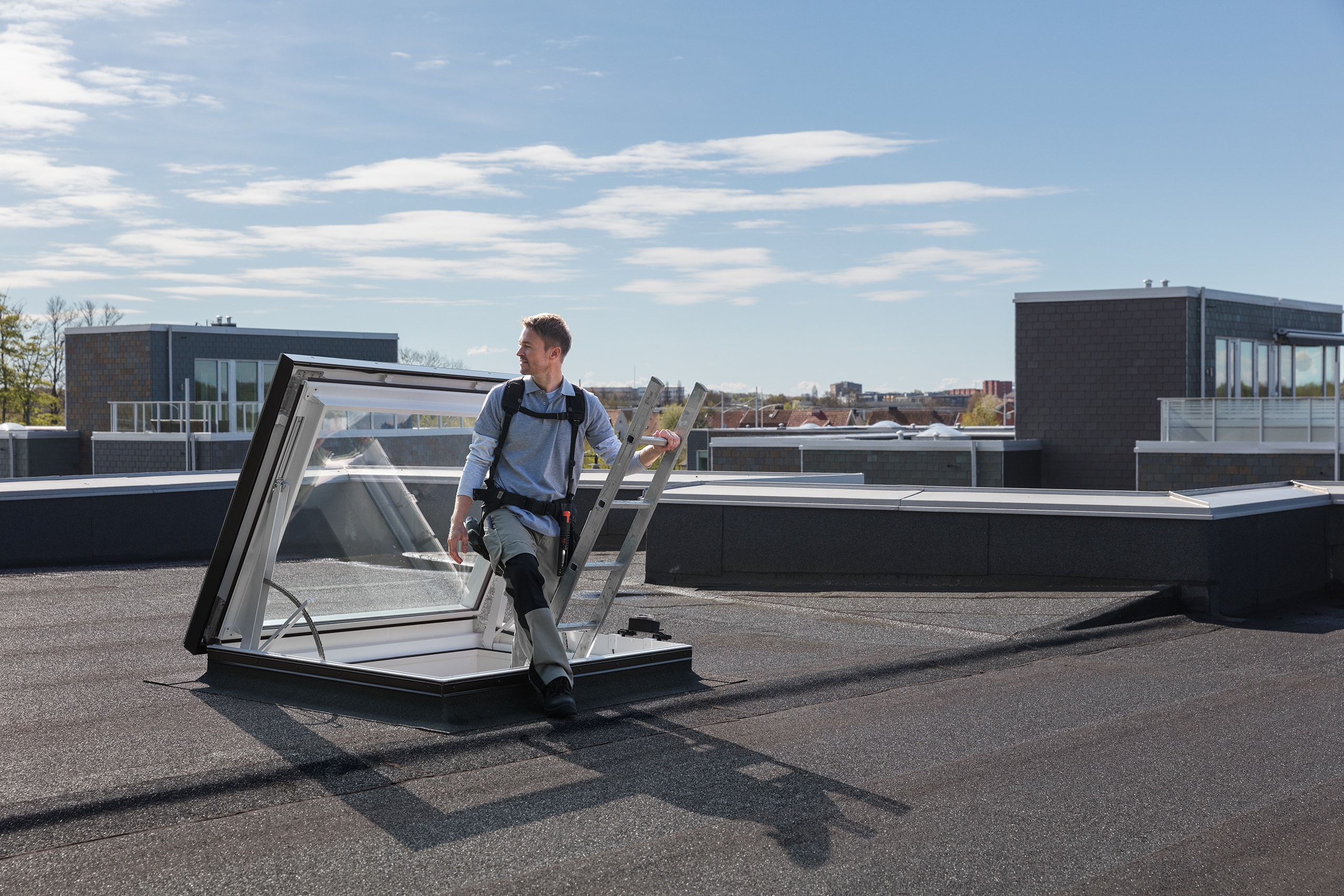 VELUX zeigt einen Techniker, der mittels Leiter durch ein geöffnetes dunkelgraues Flachglasfenster auf das Flachdach steigt.