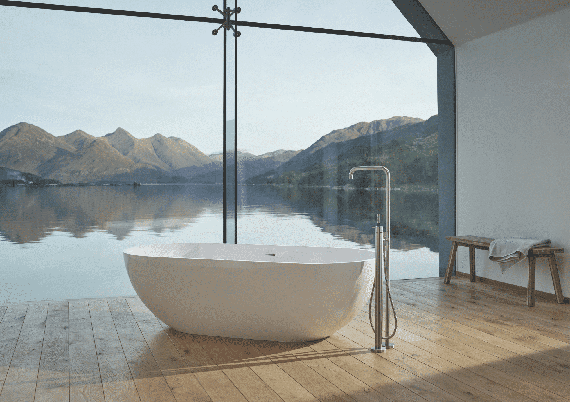 VOLA zeigt ein Badezimmer mit Holzboden und einer weissen, freistehenden Badewanne mit silbernen Armaturen und großer Glasfront mit Blick auf einen See.