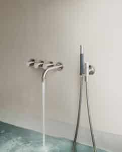 VOLA präsentiert einen Badewanneneinlauf mit drei Reglern und Handdusche in mattem Chrom in der Wand montiert.