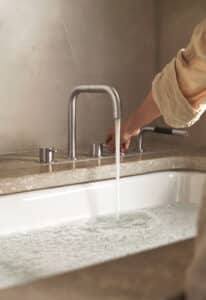 VOLA präsentiert eine Badewanne mit Wassereinlauf, drei Reglern und Handdusche in mattem Chrom am Badewannenrand.