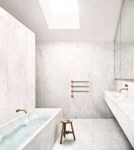 VOLA präsentiert ein Badezimmer mit Waschtisch und Badewanne aus Marmor mit Badezimmerheizung mit drei Stäben, Badewannen- und Waschbeckeneinlauf mit Eingriffmischern aus mattem Messing.