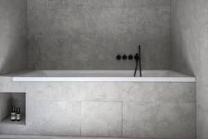 VOLA zeigt eine integrierte Badewanne mit grau marmorierten Fliesen und schwarzer Armatur mit drei seperaten Drehschaltern und stabförmiger Handdusche.