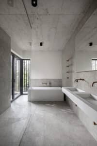 VOLA zeigt ein Badezimmer mit grau marmorierten Fliesen mit weißem Doppelwaschbecken mit wandmontiertem kupferfarbenen Wasserauslauf, Badewanne mit kupferfarbenen Armaturen und stabförmigem Handtuchhalter.