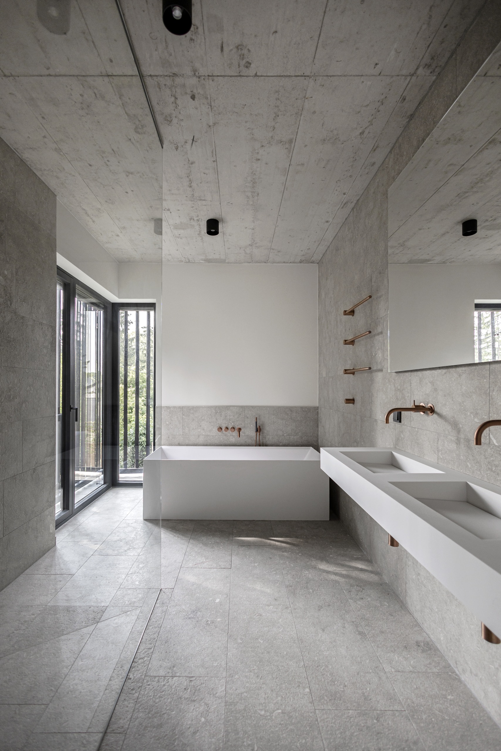 VOLA zeigt ein Badezimmer mit grau marmorierten Fliesen mit weißem Doppelwaschbecken mit wandmontiertem kupferfarbenen Wasserauslauf, Badewanne mit kupferfarbenen Armaturen und stabförmigem Handtuchhalter.