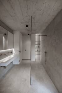 VOLA zeigt ein Badezimmer mit grau marmorierten Fliesen mit weißem Doppelwaschbecken mit wandmontiertem Wasserauslauf, stabförmiger Kopf- und Handdusche mit kupferfarbenen Armaturen.