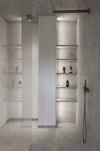 VOLA zeigt eine Dusche mit grau marmorierten Fliesen und kupferfarbenen Dusch-Armatur mit stabförmiger Kopf- und Handdusche und seperatem Drehschalter.