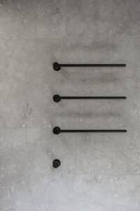 VOLA zeigt ein Badezimmer mit grau marmorierten Fliesen und einen Handtuchhalter, bestehend aus drei schwarzen Stäben.