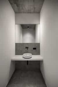 VOLA zeigt ein Badezimmer mit grau marmorierten Fliesen, weißem Waschtisch und freistehendem Waschbecken, rundem Spiegel und schwarzer Armatur.