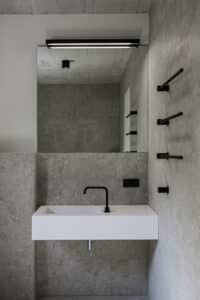 VOLA zeigt ein Badezimmer mit grau marmorierten Fliesen, weißem Waschbecken mit schwarzer Armatur und schwarzem Handtuchhalter aus drei Stäben.