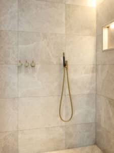 VOLA zeigt eine Dusche mit stabförmiger Handdusche in gold mit drei Drehschalten auf Marmorfliesen montiert.