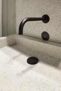 VOLA zeigt die Nahaufnahme einer Waschbecken-Armatur mit Sensor in schwarz, die in der Wand integriert ist und einer schwarzen Abflussabdeckung.