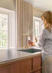 Eine Frau an einer VOLA KV5 C70 Armatur in Gold mit laufendem, dampfenden Wasser in einer modernen Küche.