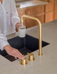 VOLA präsentiert einen Wasserhahn in gold mit zwei Drehreglern, die auf der Arbeitsplatte montiert sind, aus dem kochend heißes Wasser kommt.