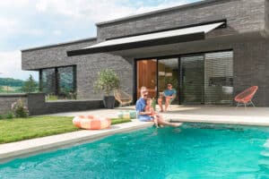 Ein Flachdachhaus mit Steinfassade, Pool, Terrasse, schwarzen Außenjalousien und Markise mit schwarzem Rahmen und hellgrauem Stoff von Warema.
