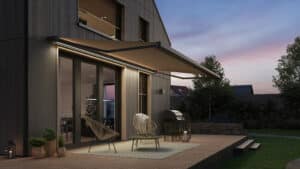 Ein Haus bei Nacht mit dunkler Holzfassade, Terrasse mit Podest und einer Markise mit dunkelgrauem Rahmen und beigem Stoff passend zu den hellen Holzfenstern und Jalousien von Warema.