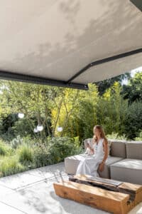 Ein Terrassenbereich mit beiger Markise in dunkelgrauem Rahmen, Outdoor‑Couch auf der eine Frau im Schatten entspannt von Warema.
