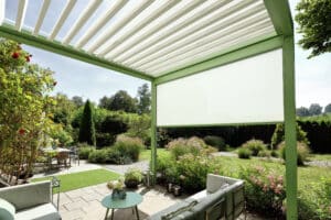 Klassische Villa mit verspieltem Garten und einer Terrasse mit Sitzgruppe unter einer grünen Pergola mit halboffenem Lamellendach und einer weissen, schattenspendenden Seitenmarkise von Warema.