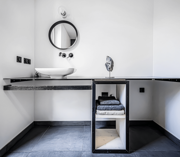 Weissenseer Holz-System-Bau zeigt modernes helles Badezimmer mit schwarzem Waschtisch, weißem freistehendem Waschbecken, schmalem Handtuchregal und schwarzen Armaturen.