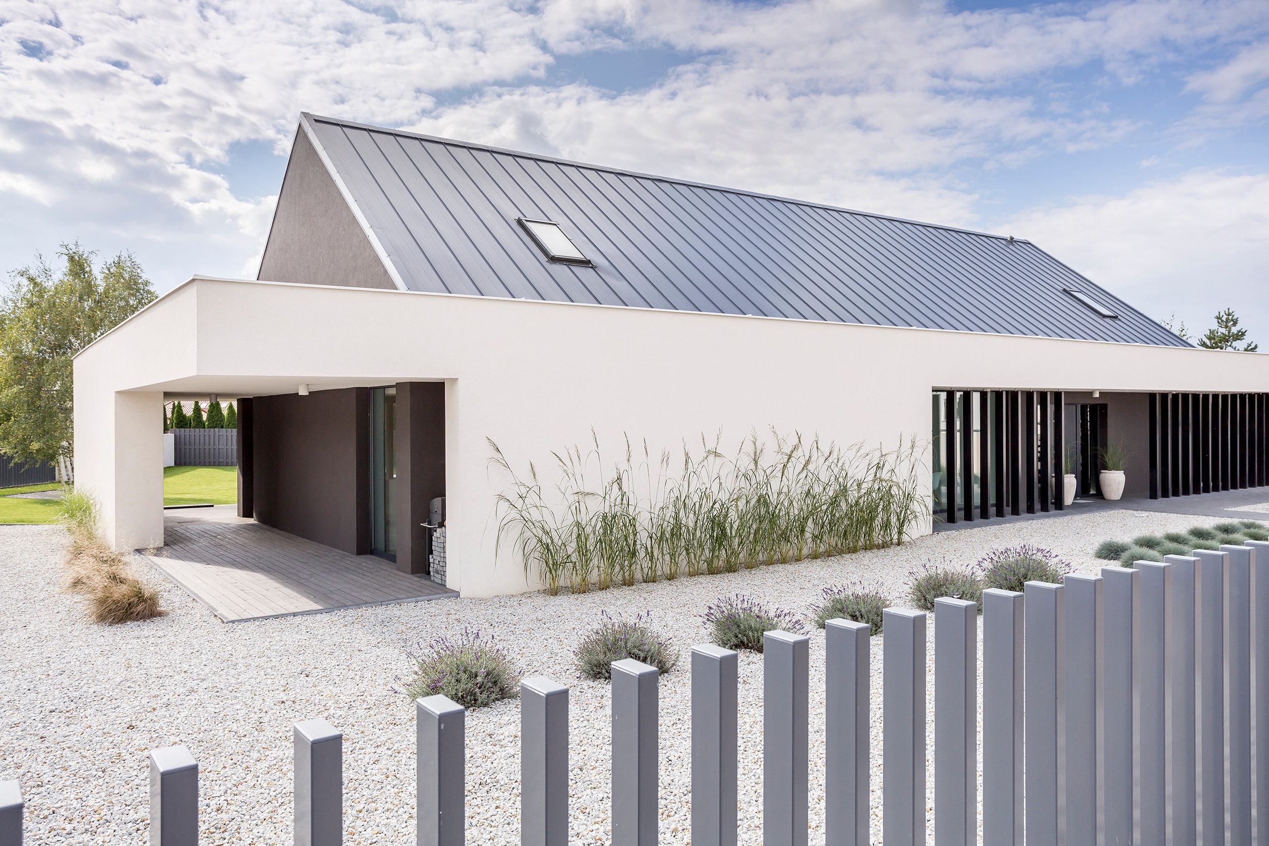 Rückansicht einer modernen, eingeschossigen Villa in weiß mit Flachdach, mit großem Kiesbereich inklusive strukturierter Bepflanzung mit Blauschwingel und Gräsern, davor ein moderner Gartenzaun mit aus dem Boden ragenden Metallstäben.