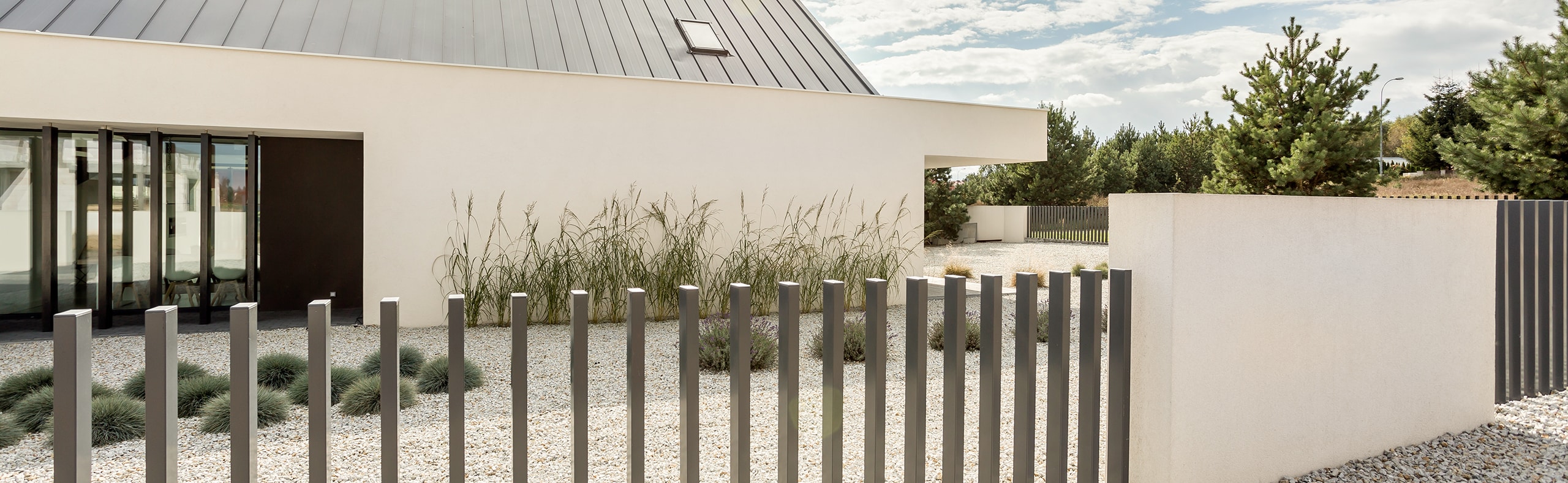 Rückansicht einer modernen, eingeschossigen Villa in weiß mit Flachdach, mit großem Kiesbereich inklusive strukturierter Bepflanzung mit Blauschwingel und Gräsern, davor ein moderner Gartenzaun mit aus dem Boden ragenden Metallstäben.