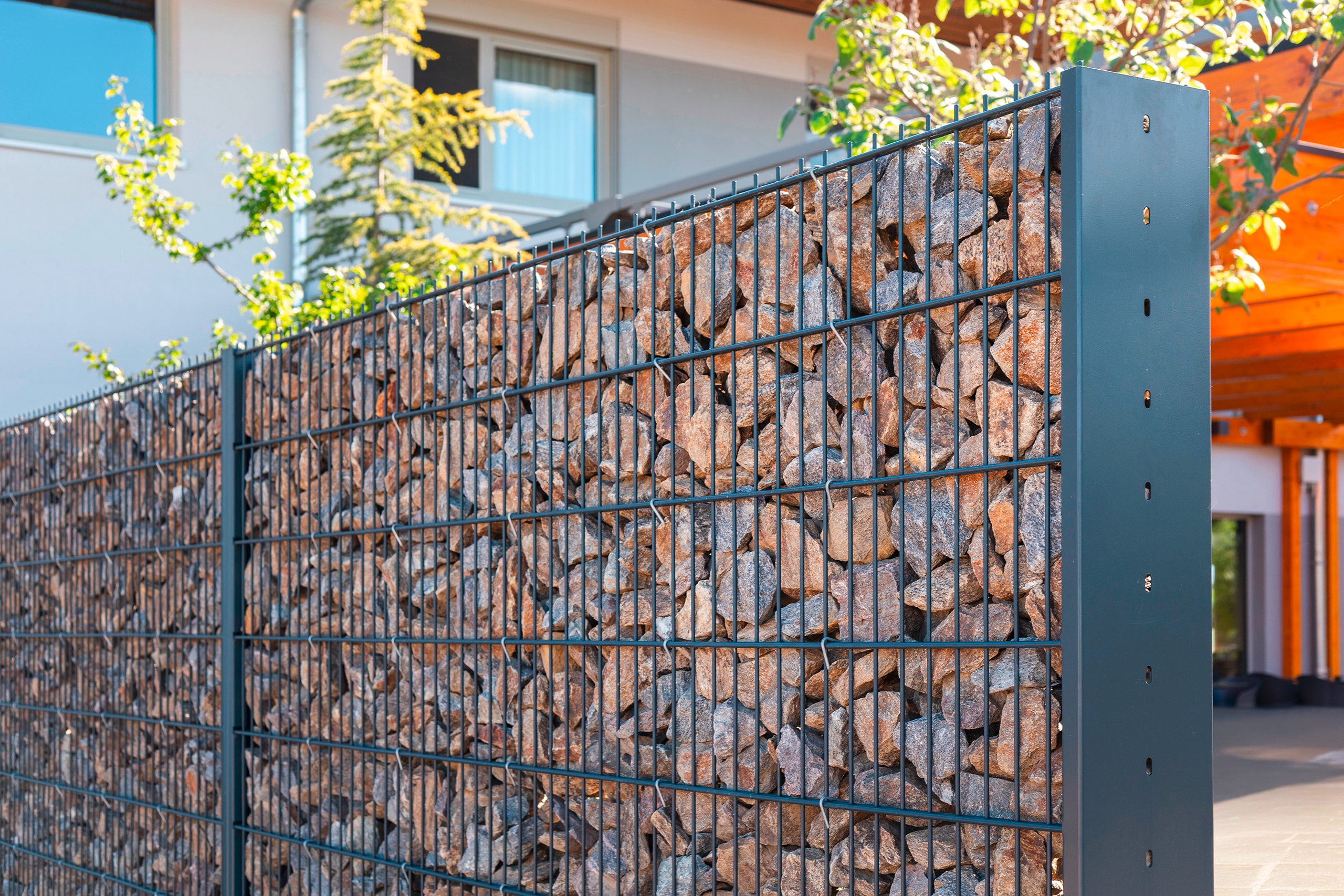 Moderner Gabionenzaun aus Steinen bildet eine elegante Abgrenzung. Er steht in einem zeitgenössischen Ambiente, ergänzt durch Grünflächen und eine elegante architektonische Kulisse, und präsentiert Innovation.