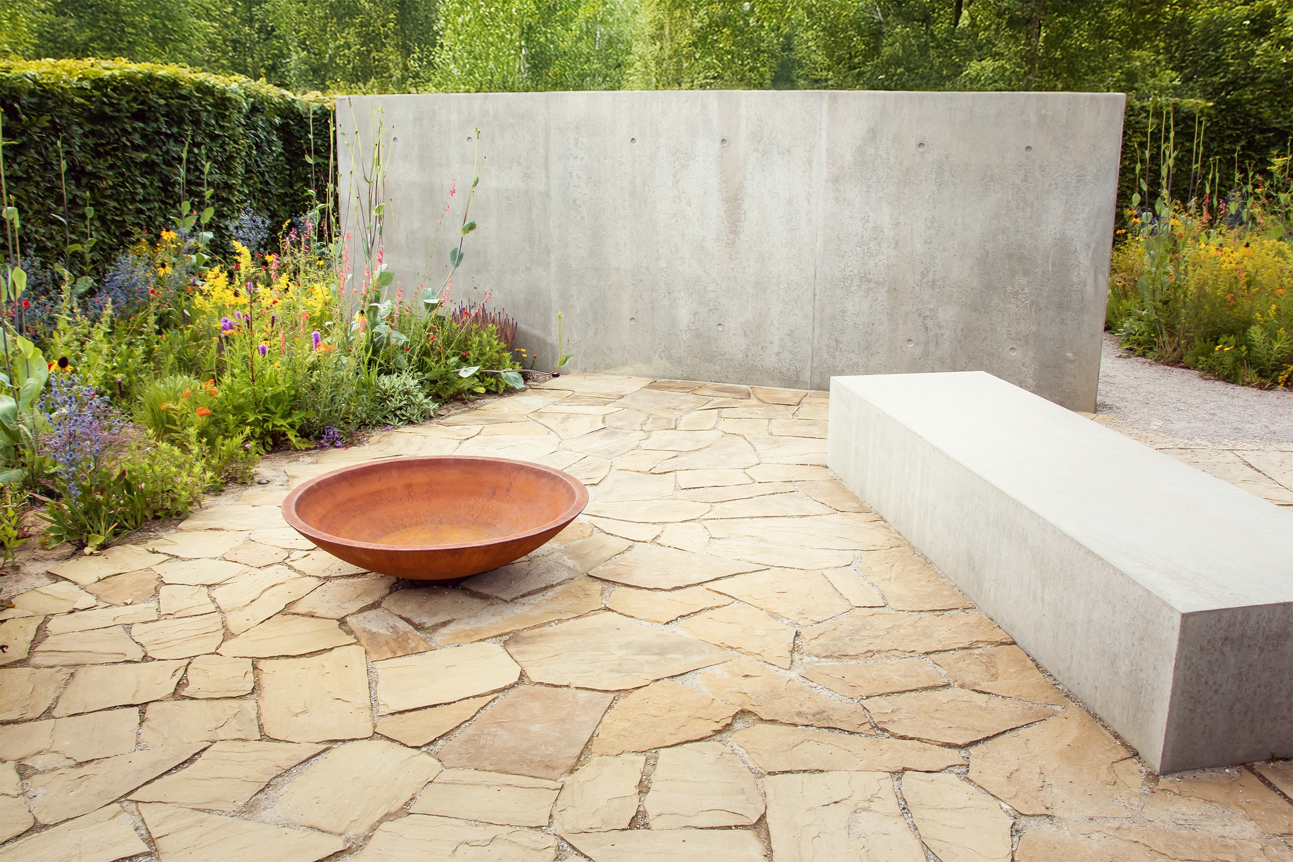 Runde Schale aus Corten-Stahl platziert am Boden aus naturgehauenen Sandsteinplatten in Mosaikverlegung inmitten eines modernen Gartens mit Betonwand und Betonblock als Sitzgelegenheit, sowie bunten Blühpflanzen.