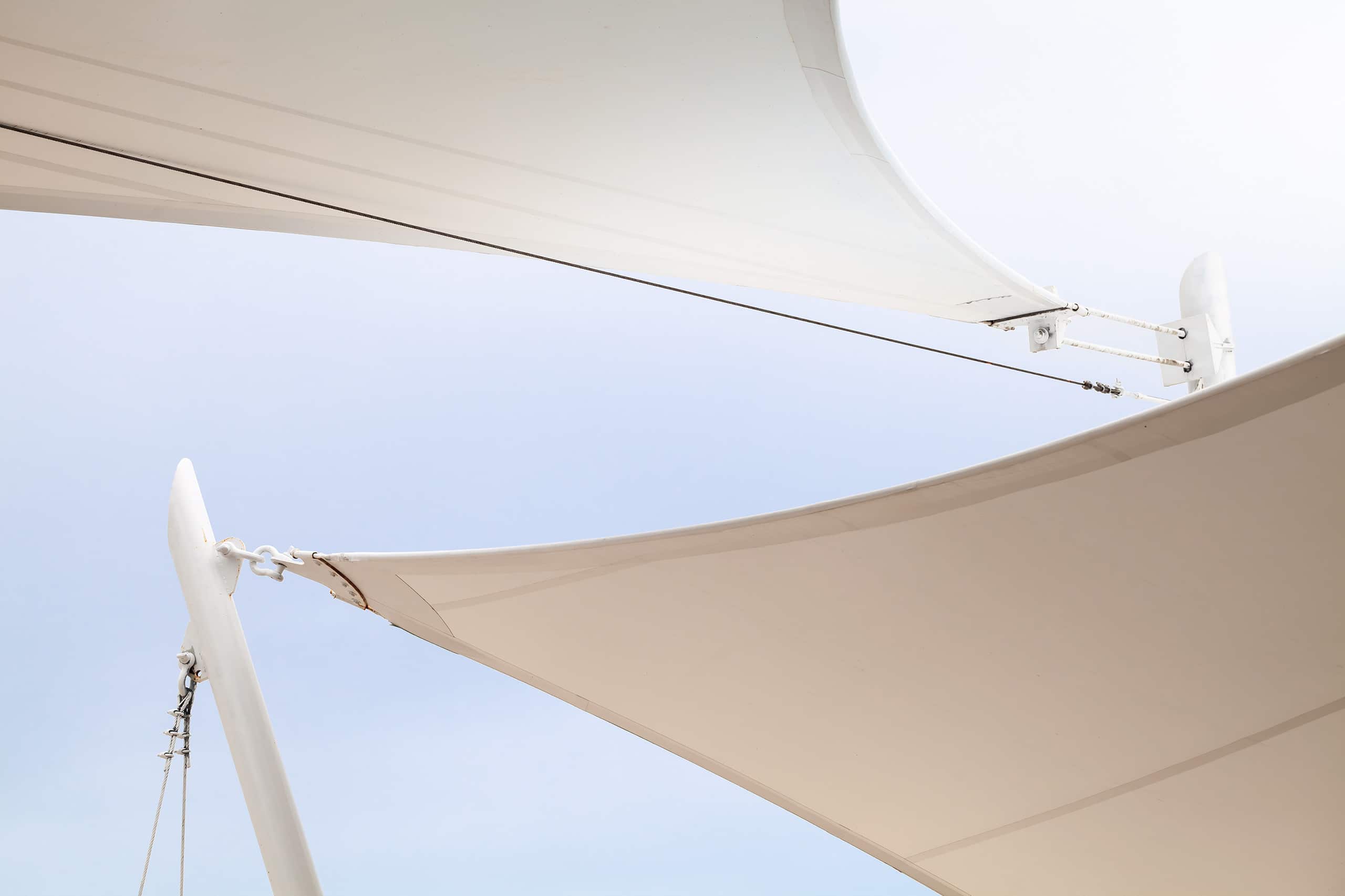Detailaufnahme eines zweiflügeligen Sonnensegels, deren Verankerung an den weißen Metallmasten gut erkennbar ist.