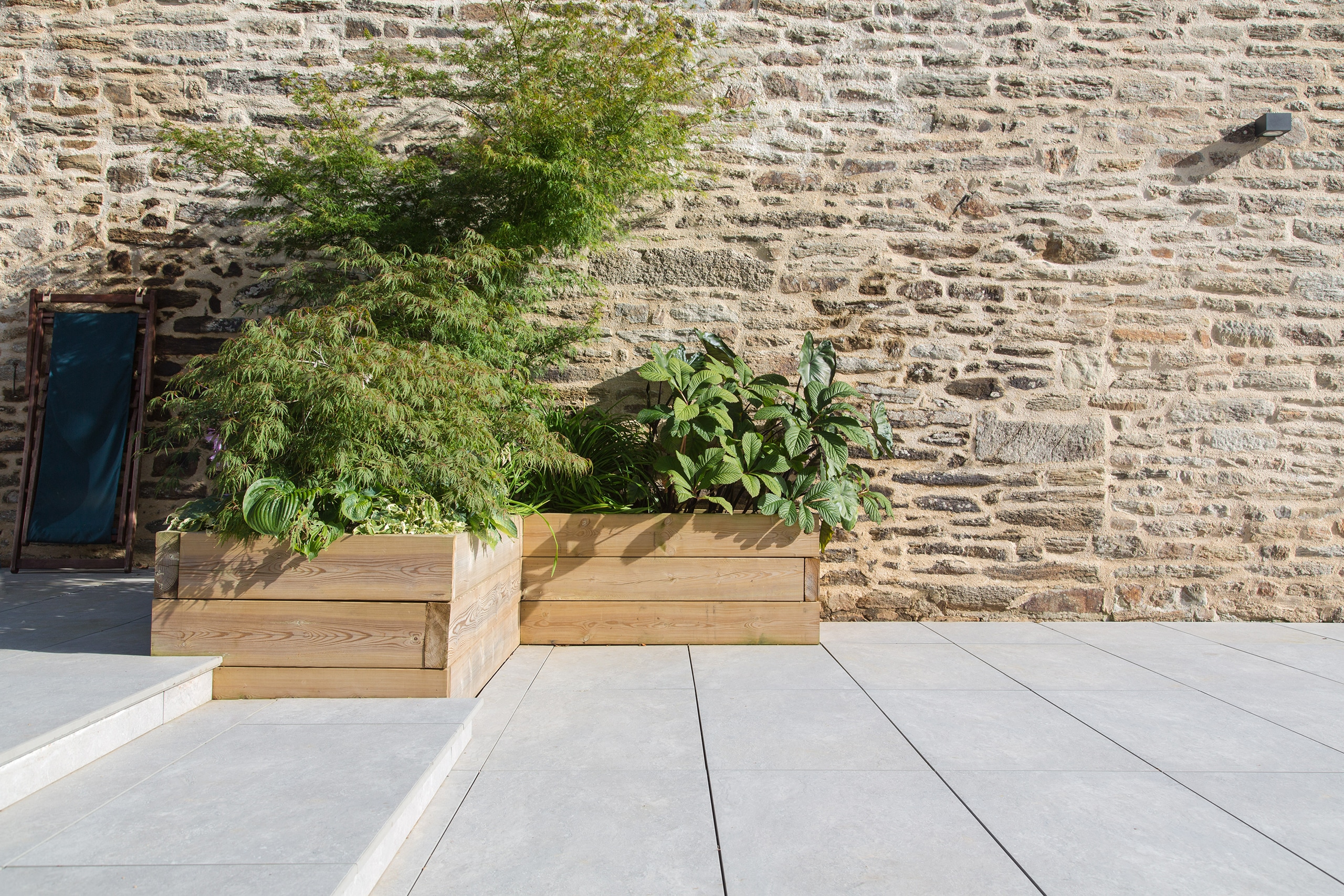 Moderne Gartengestaltung und Terrassenbau: Hanggrundstück mit Natursteinplatten im Kontrast zu Hochbeeten mit Grünpflanzen in Holztöpfen vor einer alten Steinmauer