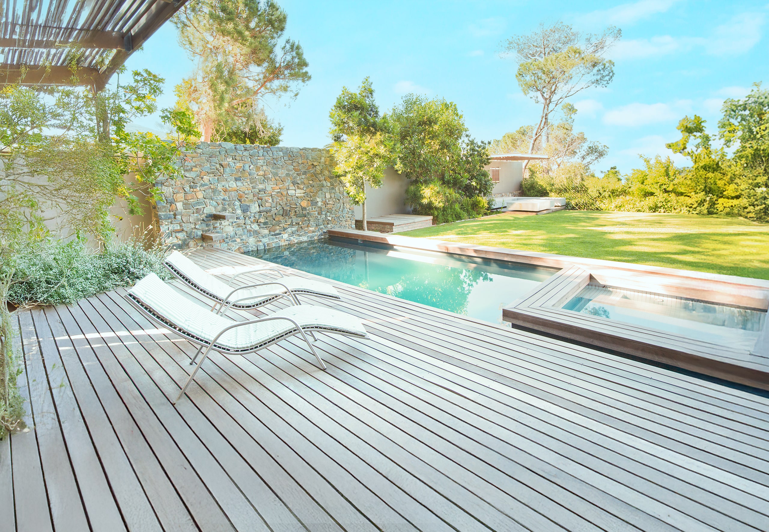 Große Terrasse mit hellen Holzdielen und Poolumrandung aus dem selben Holz und dahinter liegendem Garten mit grünem Rasen, Sträuchern und Bäumen.
