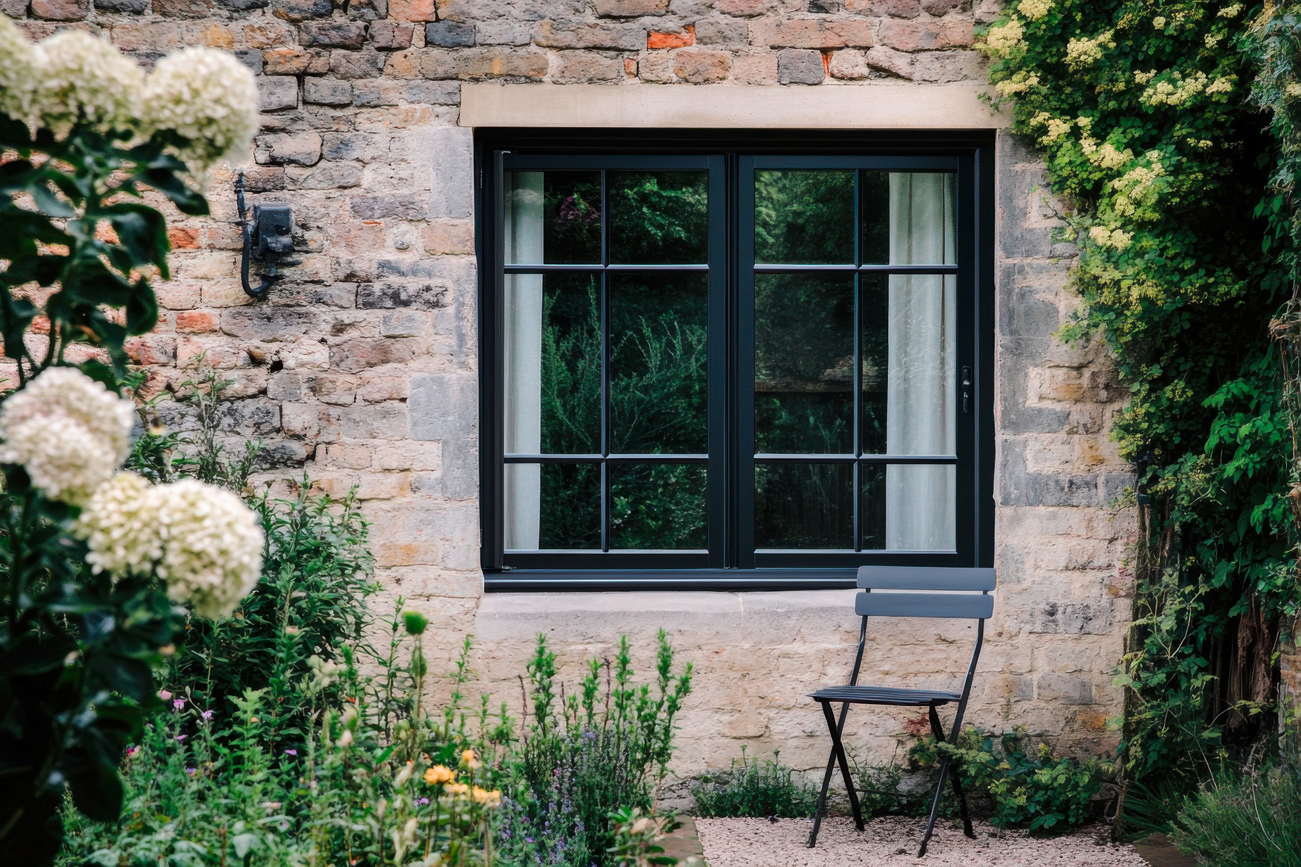 Charmante Gartenecke mit einem minimalistischen Stuhl neben einer rustikalen Steinmauer und einem großen, schwarzen Sprossenfenster mit Blick ins Grüne in ruhiger Lage.