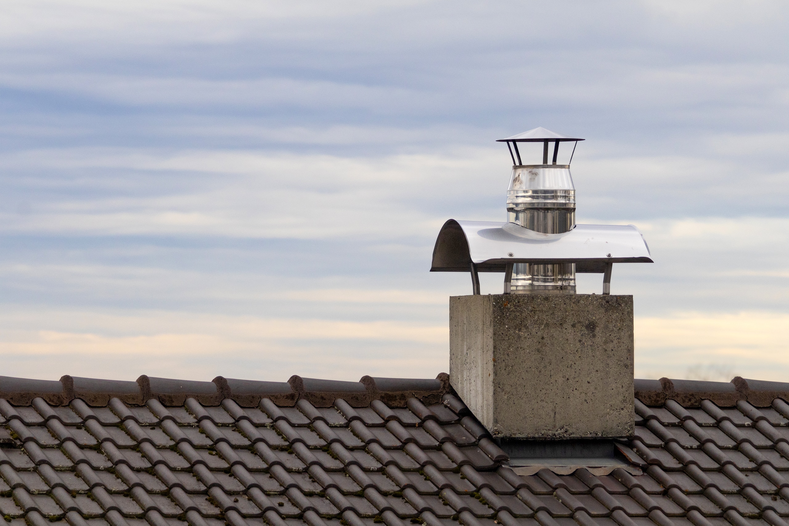 Betonierter, kubusförmiger Rauchfang (Schornstein) mit Edelstalrohr auf dem Dach eines Einfamilienhauses mit dunkler Dacheindeckung bei Abendstimmung.