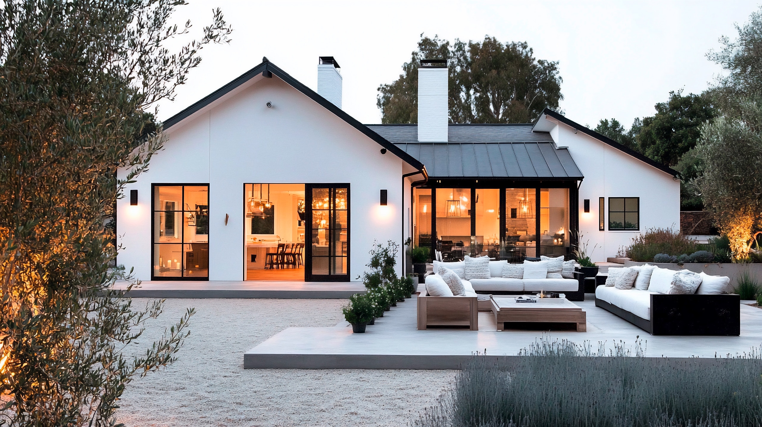 Außenansicht eines modernen weißen Hauses mit markanten, schwarzen Leisten im Dachbereich, großen Glasfenstern und Gartenmöbeln. Das Haus wird von Innen- und Außenleuchten beleuchtet und strahlt ein warmes Licht aus. Ein Kieshof und die Gartengestaltung runden den Außenbereich ab.