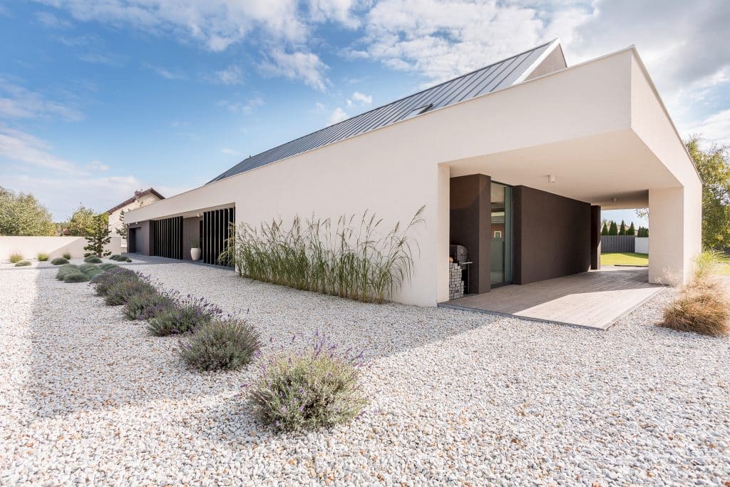 Moderne, eingeschossige Villa in weiß mit Flachdach, vertikalen Designelementen aus dunklem Holz und überdachter Terrasse mit Blick auf das große Kiesbeet mit strukturiert gepflanzten Gräsern und Lavendel..