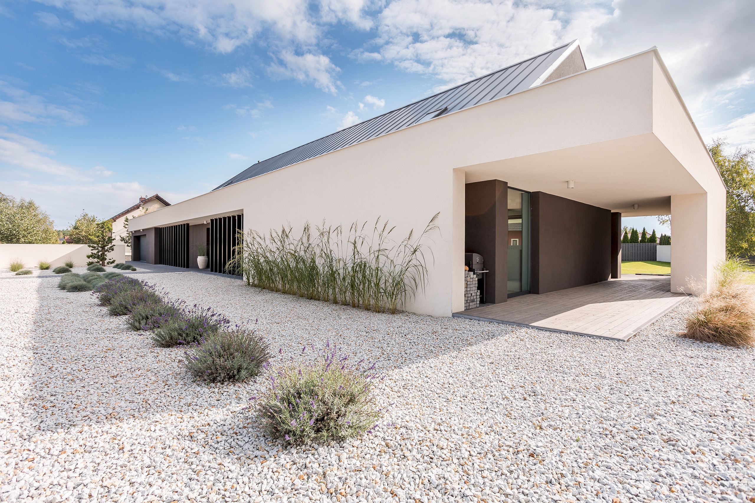 Moderne, eingeschossige Villa in weiß mit Flachdach, vertikalen Designelementen aus dunklem Holz und überdachter Terrasse mit Blick auf das große Kiesbeet mit strukturiert gepflanzten Gräsern und Lavendel..