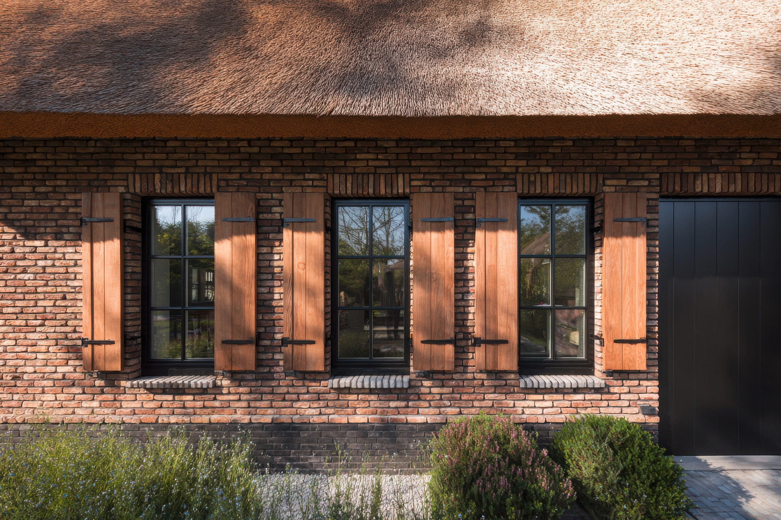 Ansprechende rchitektur verschmilzt mit der Natur in einem charmanten Backsteinhaus mit Reetdach und großen, eleganten Fenstern, das von Grün umgeben ist.