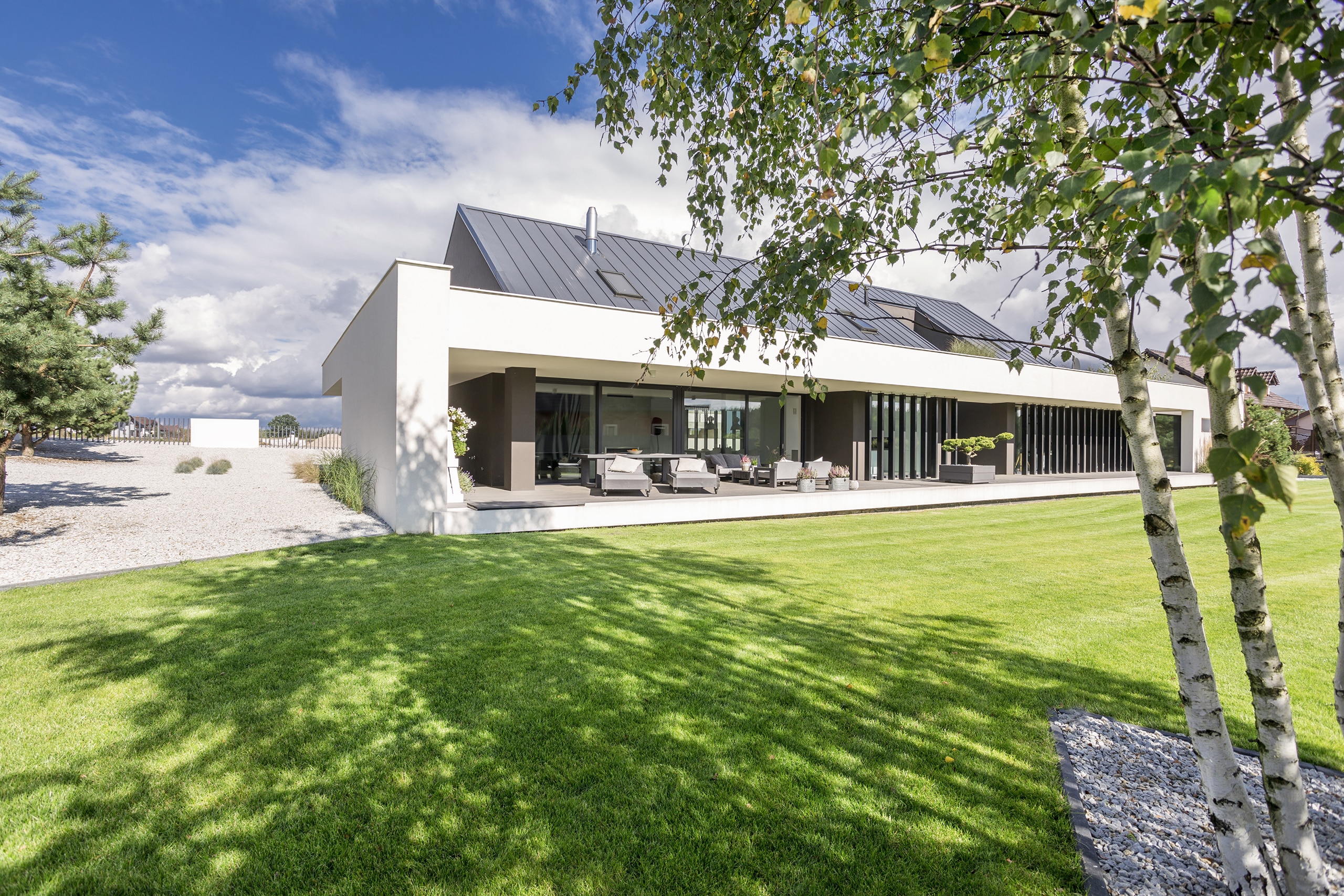 Moderne, eingeschossige Villa in weiß mit Flachdach, vertikalen Designelementen aus dunklem Holz und großzügiger Terrasse mit Lounge-Gartenmöbel mit Blick auf den frisch gemähten Rasen.